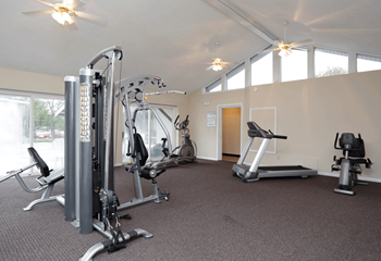 Fitness center with 4 pieces of fitness equipment, peaked ceiling and windows along the back wall.  Brown carpet.